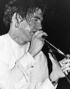 A person with wet hair, wearing a white shirt and jewelry, sings into a microphone on stage under bright lights.